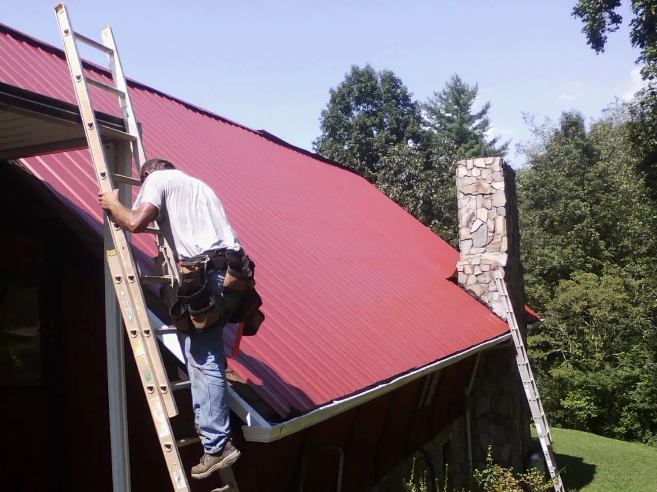 Roofer on a ladder beside a red metal roof and stone chimney — Soffit & Fascia Repair by Roofing Brothers in Elk Grove Village